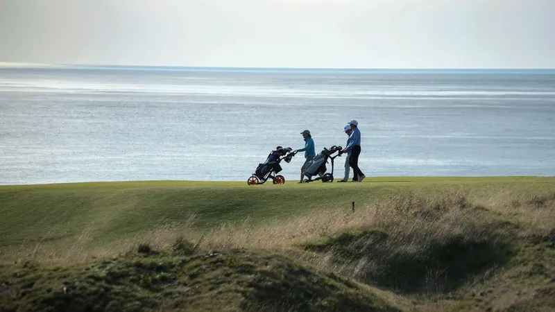 Golfers pulling trolleys along a coastal fairway
