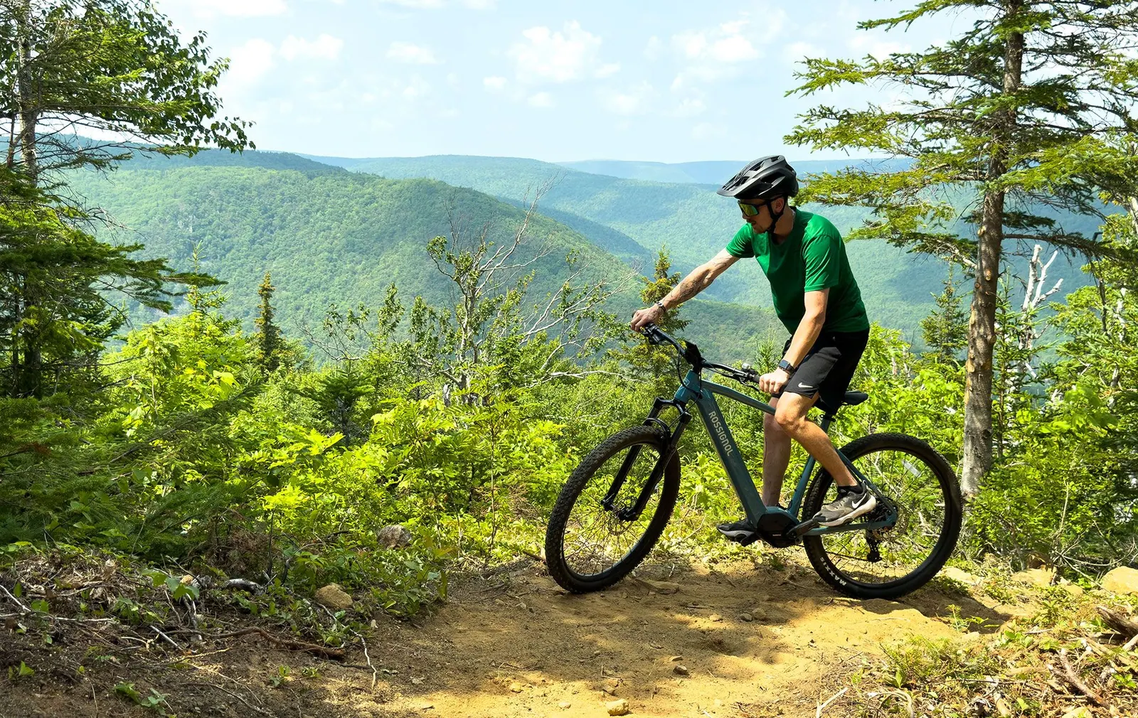 Two riders on e-bikes on a dirt trail high above the green Cape Breton Highlands