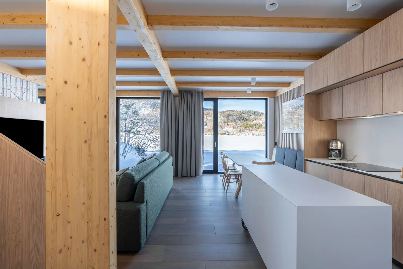 Chalet interior with timber beams, a long white kitchen island and windows onto the snowy bay