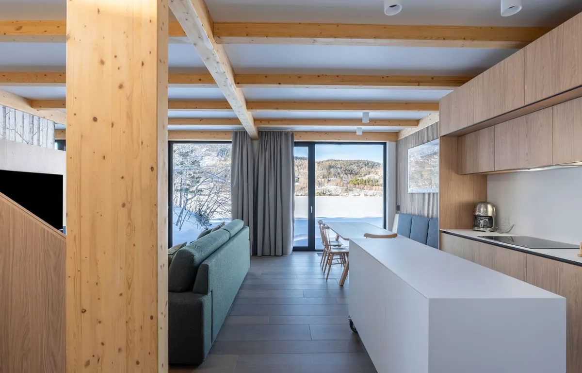 Chalet interior with timber beams, a long white kitchen island and windows onto the snowy bay