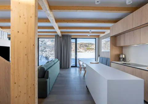 Chalet interior with timber beams, a long white kitchen island and windows onto the snowy bay