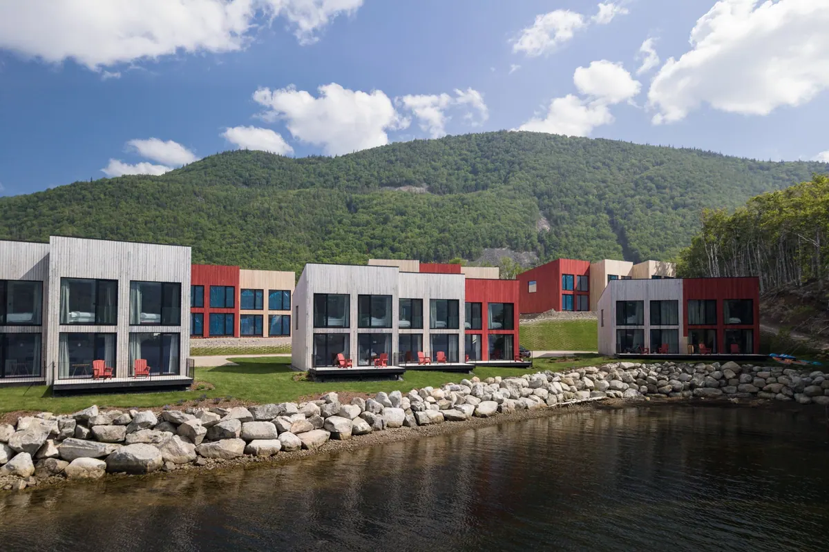 Row of white, grey and red chalets above a stone seawall, beneath Cape Smokey on a summer day