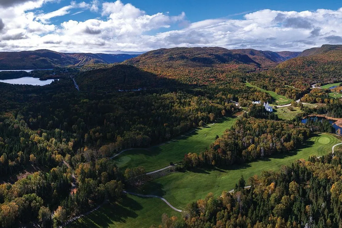 Aerial view of golf fairways winding through forested hills with lakes and mountains beyond