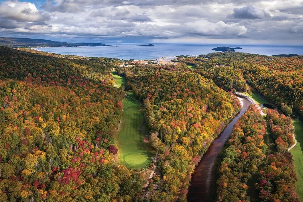 Aerial view of a fairway cut through autumn forest beside a river, with the ocean and islands behind