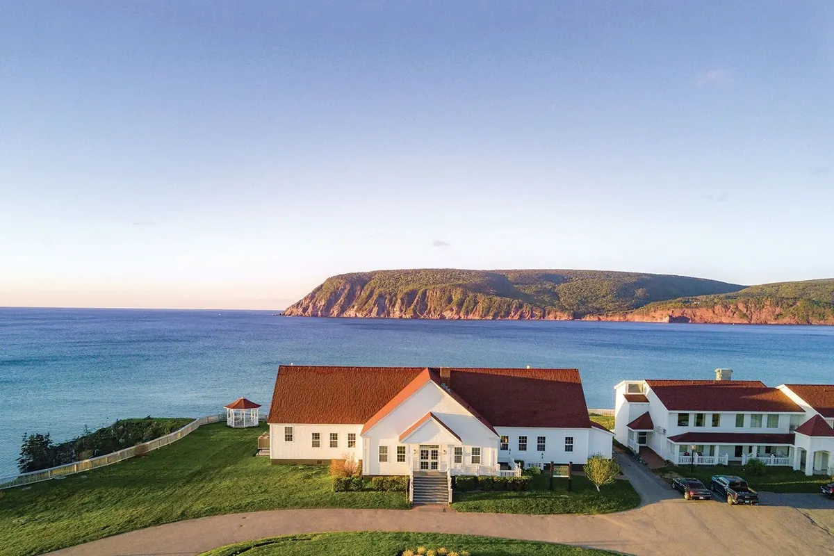 White lodge with a red roof on a grassy headland above the Atlantic, with cliffs across the bay