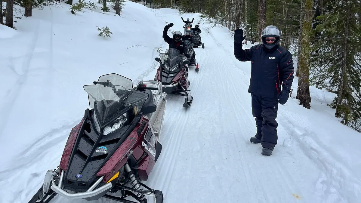 Snowmobiler waving beside parked sleds on a snowy trail