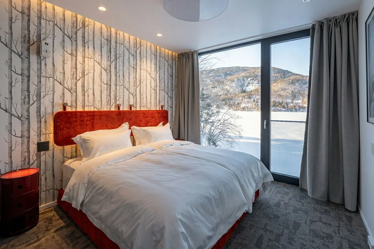 Chalet bedroom with a red headboard, birch-tree wallpaper and floor-to-ceiling windows onto the winter bay