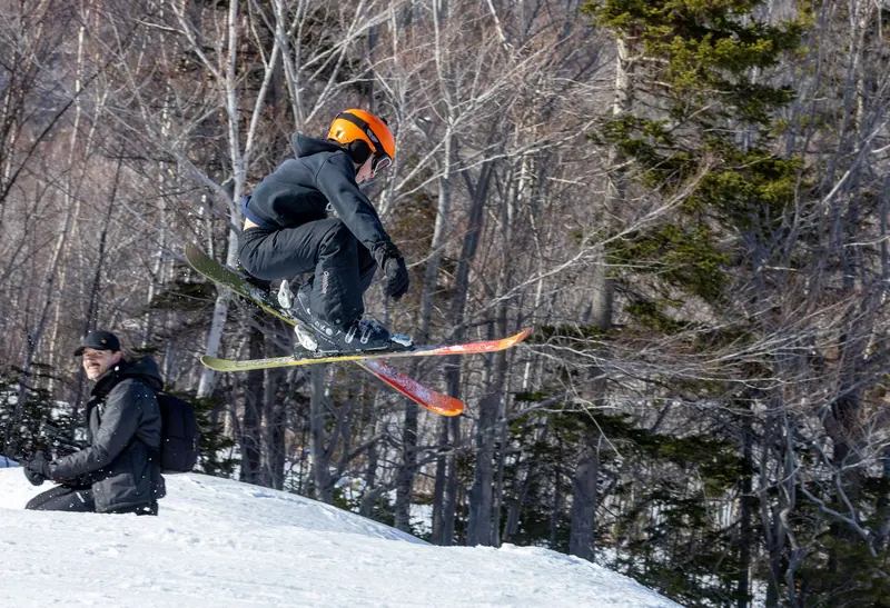 Skier jumping between birch trees while a photographer watches